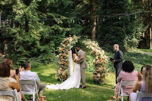 Wedding ceremony in forest setting, couple kisses in front of floral arch.