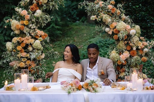 Wedding couple sit at decorated table framed in background by floral arch